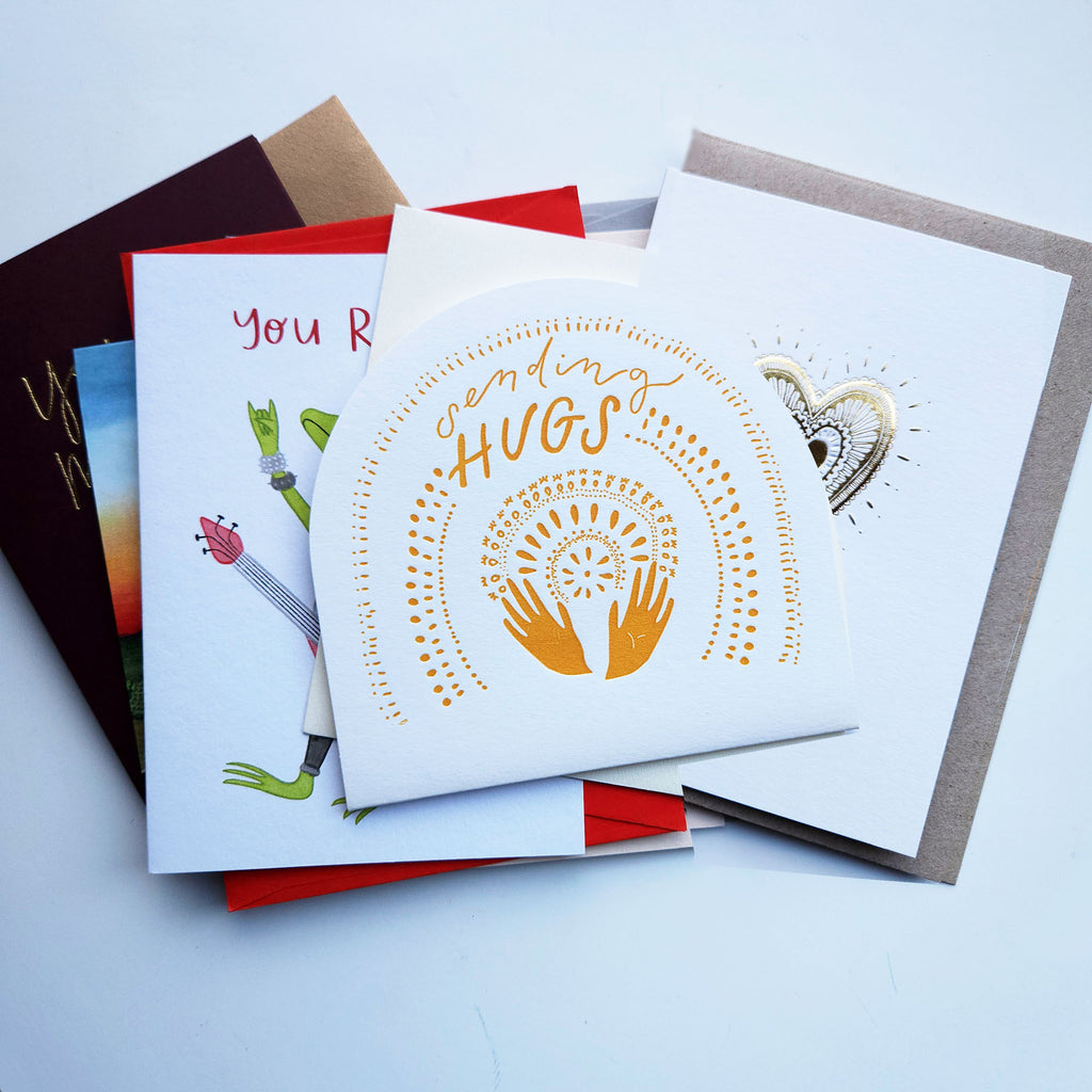 A stack of Australian greeting cards and envelopes. The top card is white with gold foil text reading 'sending hugs' and a gold heart-and-hands design.