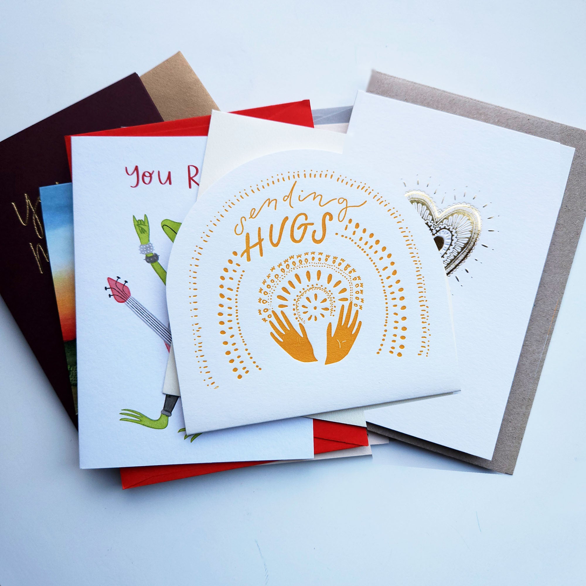 A stack of Australian greeting cards and envelopes. The top card is white with gold foil text reading 'sending hugs' and a gold heart-and-hands design.