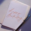 A white textured greeting card with elegant cursive script reading 'happy birthday,' shown with a cream envelope.