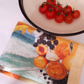 Tea towel with a colorful fruit and landscape print beside a white bowl containing cherry tomatoes on the vine.