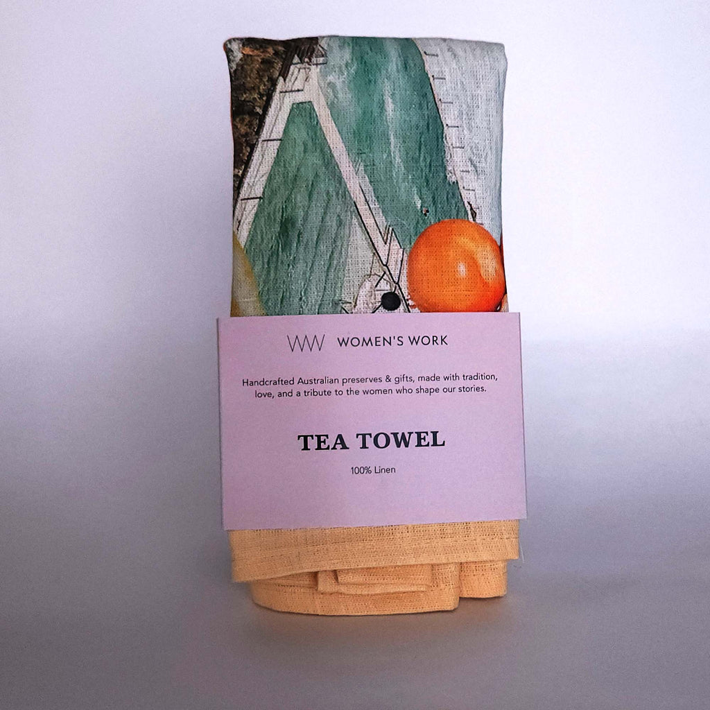 Folded tea towel with a colorful print wrapped with a paper label that reads 'WW WOMEN'S WORK TEA TOWEL 100% Linen'.