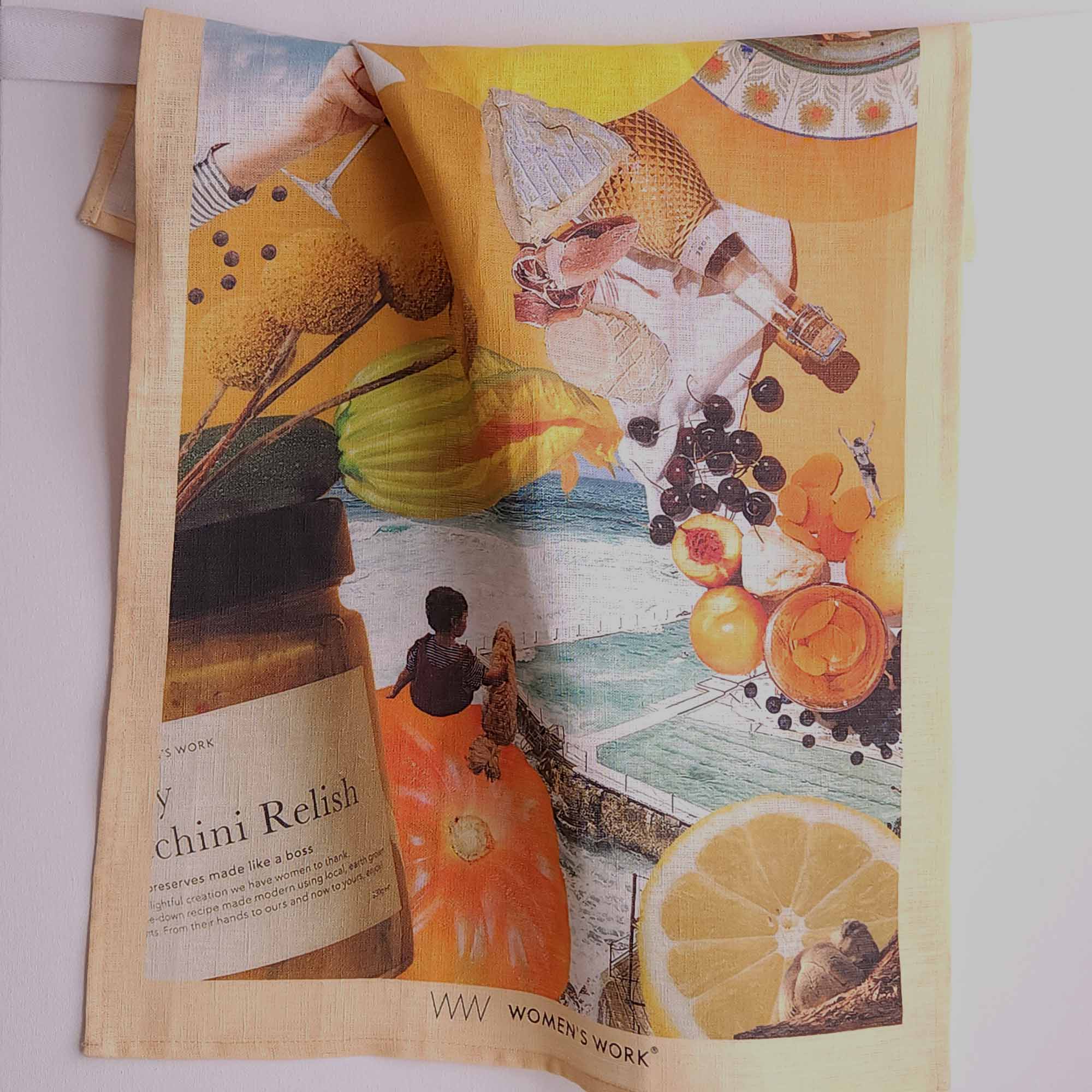 Linen tea towel with a colourful vintage-style collage with fruit, wine, and a seaside scene. The towel has a sand coloured border with 'Women's Work' branding printed near the bottom.