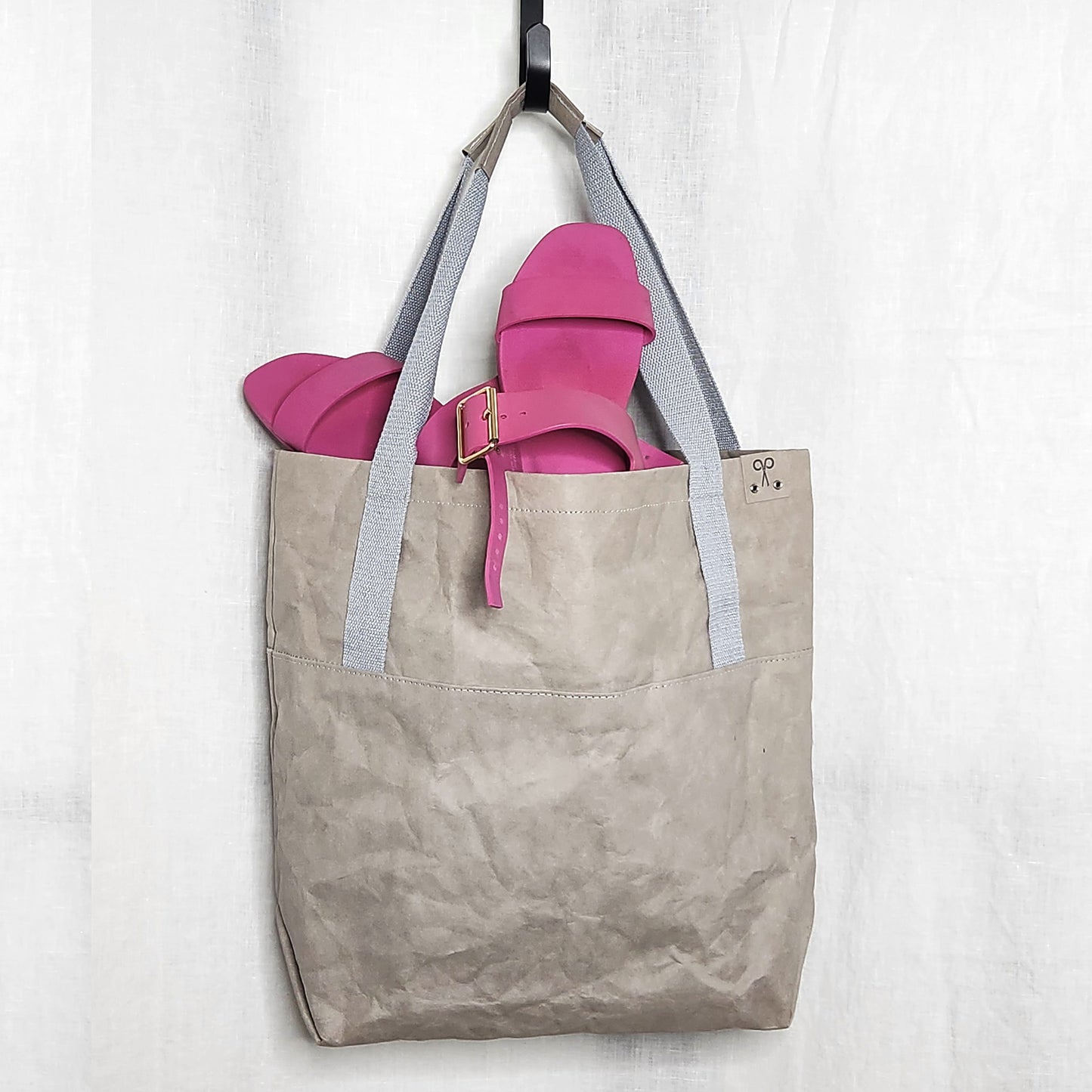 Grey washable paper tote bag with pink sandals hanging on a hook against a white background.
